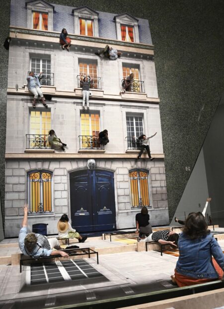 A photograph of an installation by Leandro Erlich. The installation is of the facade of a multistory home which is placed on the ground. Visitors position themselves in precarious situations, like holding onto a second-story railing and the image is brought to life using a large mirrored surface which reflects the scene making it appear as visitors are actually in danger. 