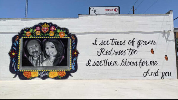 A photograph of a mural of Irma and Joe Garcia. A black and white image of the couple is framed by colorful flowers. To the right of the image are the words, "I see trees of green, red roses too, I see them bloom for me and you."