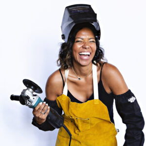 A photograph of Jessica Bell. She wears a yellow apron and holds an electric tool in one hand while wearing a welding helmet that is lifted to reveal her face. She appears to be in mid-laugh with a wide smile.