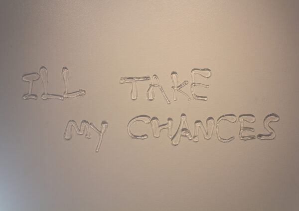 The words "ILL TAKE MY CHANCES" formed by hand ladled glass in messy font are installed onto a white gallery wall with pins. The glass is clear and contains no color.