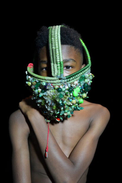 A photograph of a young Black boy wearing a sculptural piece that fits on his head and covers his nose and mouth. The piece is mostly shades of green and made with a variety of found objects and beads.