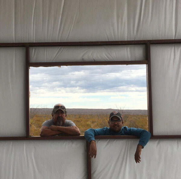 A photograph of artist Ramon G. Deanda (left) and his father (right). The two pose through an open window of a building still under construction.