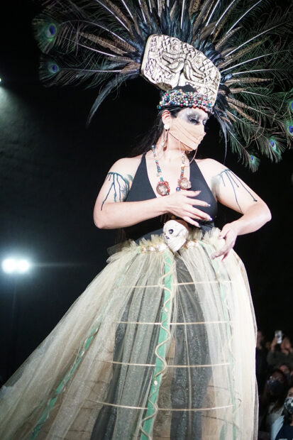 A photograph of a runway model wearing a large headdress made from peacock feathers, a black halter top, and a hooped skirt.
