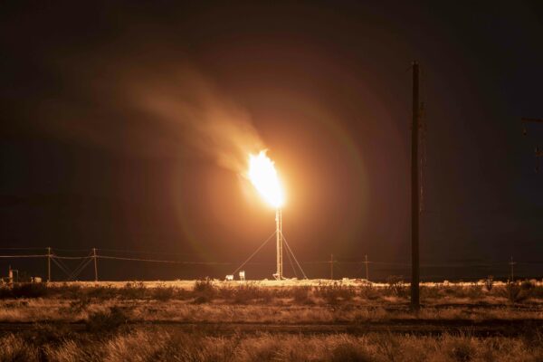 En esta fotografía una antorcha de gas ilumina el cielo nocturno y los arbustos desérticos que la rodean. En primer plano una vía corre paralela a las líneas eléctricas en el fondo.