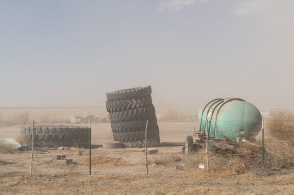 Fotografía de un paisaje desértico. Detrás de una cerca de alambre, entre varios objetos cubiertos de arena, se distinguen una torre de llantas de tractor al lado de otra llanta gigantesca y la parte cilíndrica de un camión cisterna pequeño.