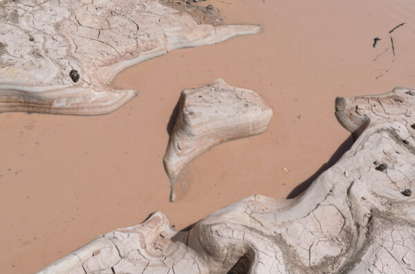 Fotografía abstracta de un paisaje lodoso que podría haberse tomado de lejos o de cerca. En ciertas partes el lodo parece ser líquido y ha adquirido un color café pálido, casi rosado, y en otras el lodo se ha secado en capas blancas y rosadas, creando formaciones geográficas redondeadas que parecen emerger de un río turbio.