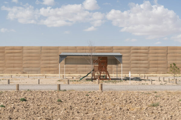 Fotografía de un pequeño parque infantil solitario bajo una estructura de metal. Detrás hay un muro alto cubierto de tela café.