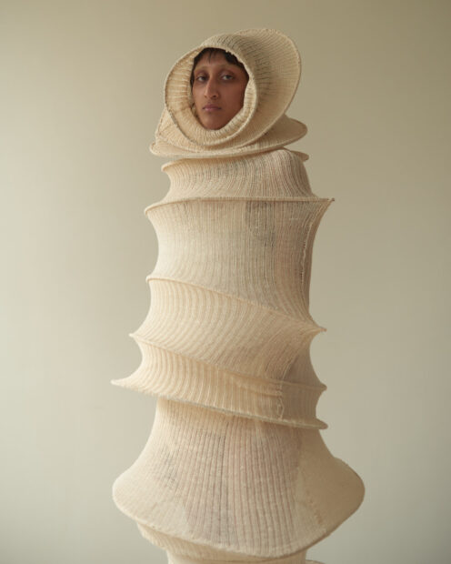 A photograph of a person inside a fiber sculpture made from white cotton and bamboo. The sculptural piece consists of about a dozen hoops spaced apart with fabric stretched across them. The form is reminiscent of a a cocoon. 