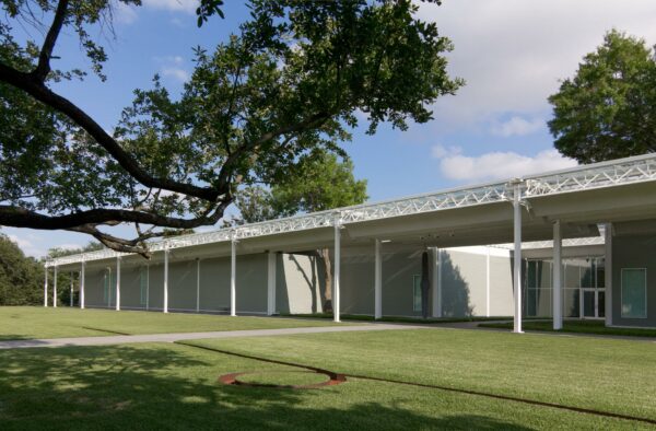 A photograph of the exterior of the Menil Collection which is a wide single story building with a sprawling lawn and large trees.