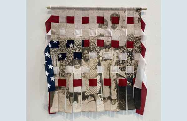 A textile work by Tina Medina. The work includes a photograph printed on fabric and strips of a cut U.S. flag. The photograph has been cut vertically into strip and the flag has been woven horizontally into the image.