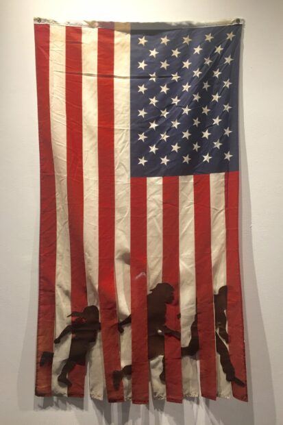 A fabric work by Tina Medina using the US flag with silhouetted figures placed between the stripes.