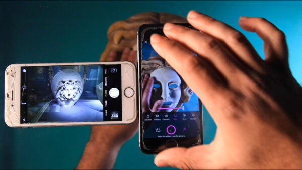 A photograph of hands touching two phones. The phones appear to be photographing artifacts and masks. The scene feels very mystical.