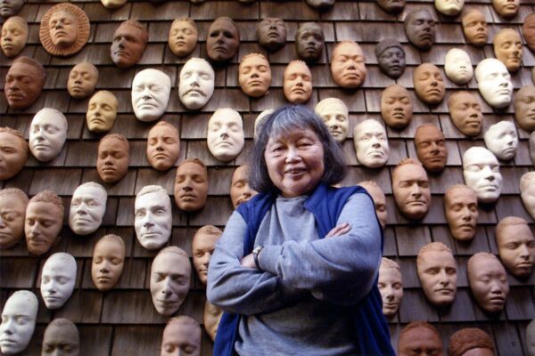 A photograph of artist Ruth Asawa standing in front of an exterior wall of her house with dozens of clay masks hanging on the wall. She stands with her arms crossed and looks at the camera with a smile.