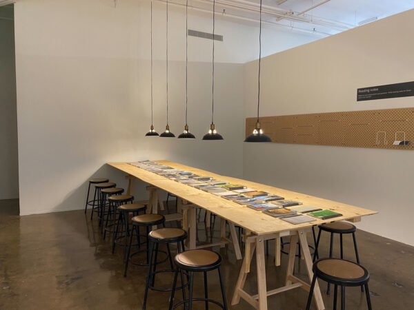 Installation view of books lined up on a natural wood table with stools surrounding