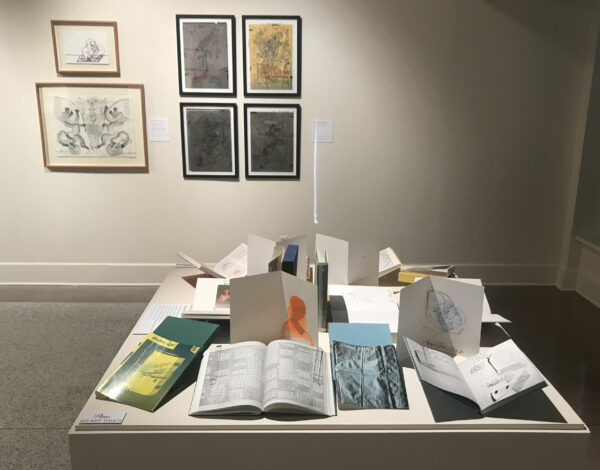 Installation view of a pile of books on a pedestal and works on paper in the background