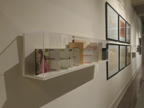 Installation view of books in a case and works on paper framed in a grid