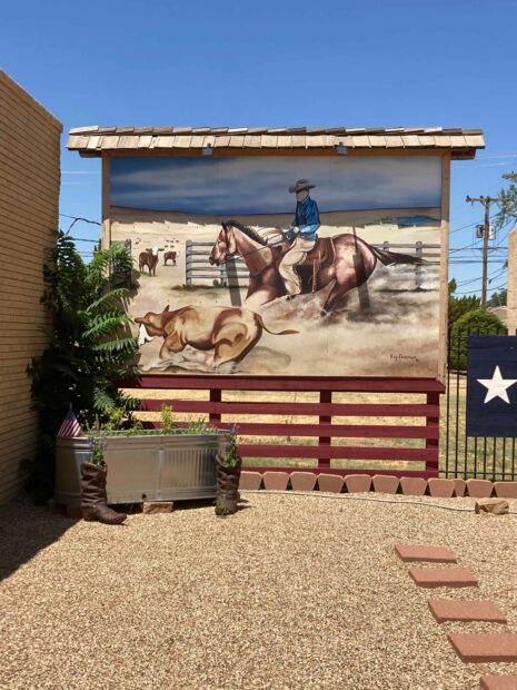 A photograph of a mural, painted on a standing billboard. The painting depicts a cowboy on a horse in a pen, riding next to a running cow. 