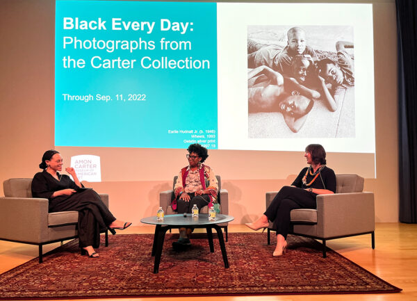 Tres mujeres están sentadas en sillones individuales en frente de una proyección que dice “Black Every Day: Photographs from the Carter Collection” [Negro todos los días: Fotografías de la colección Carter]. 