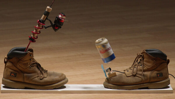 An artwork featuring a pair of boots. In one shoe is a fishing pole. Attached to the other shoe is a can of beer.
