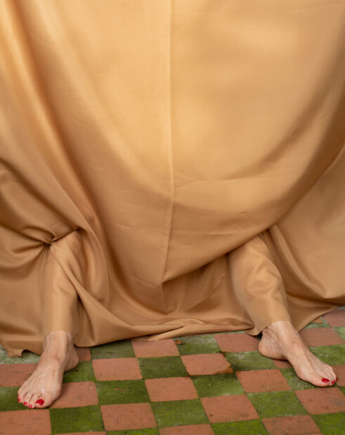 A photograph by Cristina Velásquez of a person behind a tan colored fabric. The person's feet stick out from under the fabric and rest on a patterned floor of green and brown squares.