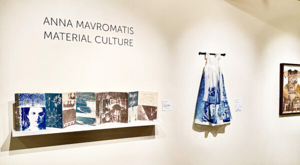 Installation view with an accordion style art book on a shelf, a girl's dress hanging on a rod, and a framed two dimensional work