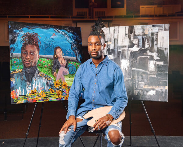 A photograph of artist Randy Palmer sitting in front of two of his paintings.