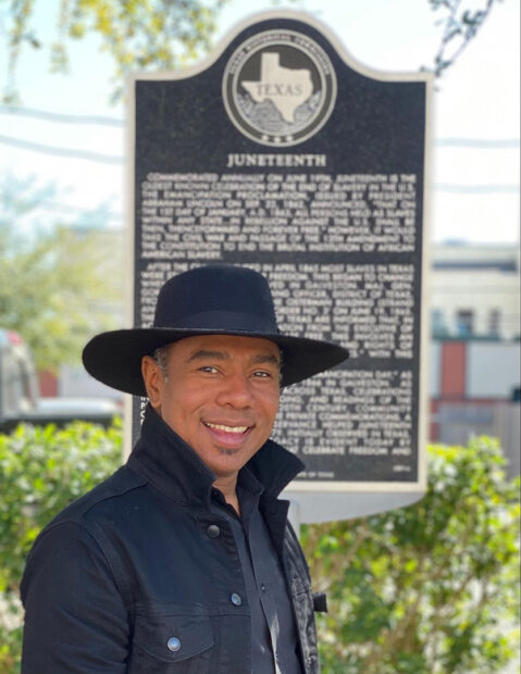A photograph of artist Reginald C. Adams standing in front of a historical placard about Juneteenth.
