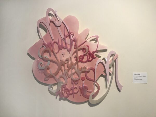 Sculpture of letters with a pink backing hanging on a wall