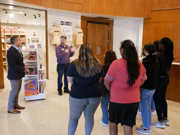 Un grupo de seis mujeres jóvenes está de pie y parece escuchar a un hombre adulto frente a la tienda de regalos de un museo.