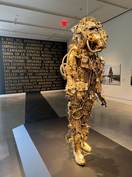 Installation view of an gold astronaut sculpture with a black and gold words in the background