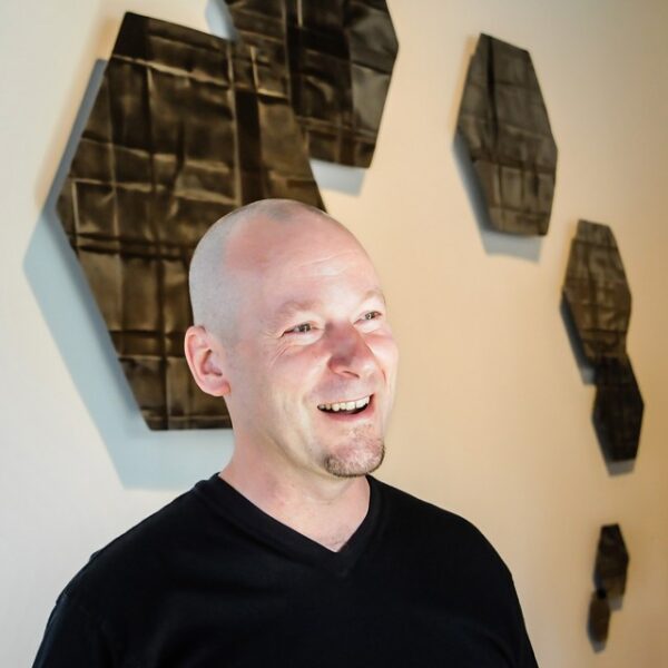A photograph of Devon Christopher Moore standing in front of a work of art and smiling as he looks off camera.