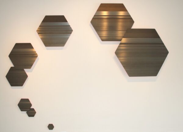 A photograph of a multi-piece steel work by Devon Moore. The artwork includes eight hexagonal sheets of metal. The sheets are installed on the wall to create a spiral. Each sheet is smaller than the previous one and has horizontal scoring marks across 75% of the sheet.