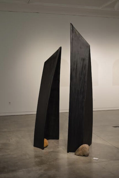 En una sala de museo con paredes blancas, se alza una escultura formada por las dos partes asimétricas de una barca negra acomodadas de manera vertical. Cada parte tiene una roca en su base.