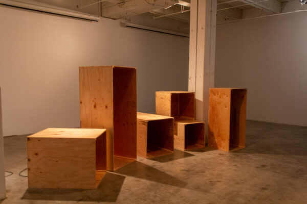 Six wooden boxes with one open face sit positioned towards the center of an empty white-walled gallery.