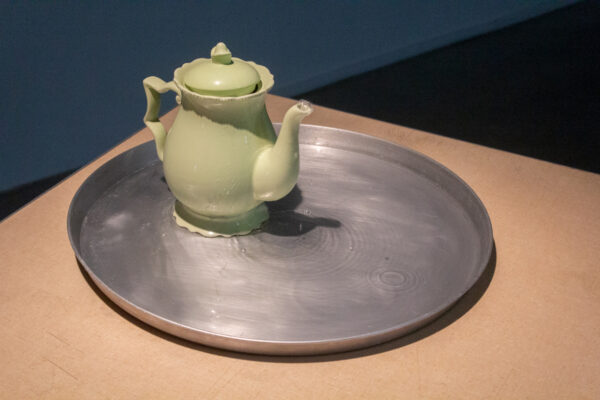 A green decorative teapos consistently bubbles water out of its spout onto a flat shallow metal pan on a tan table.