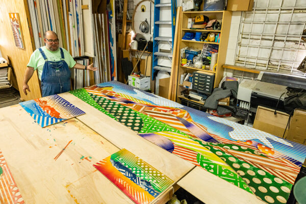 A photograph of artist Zeke Williams in his studio with a large work in progress laid out on the table in front of him.
