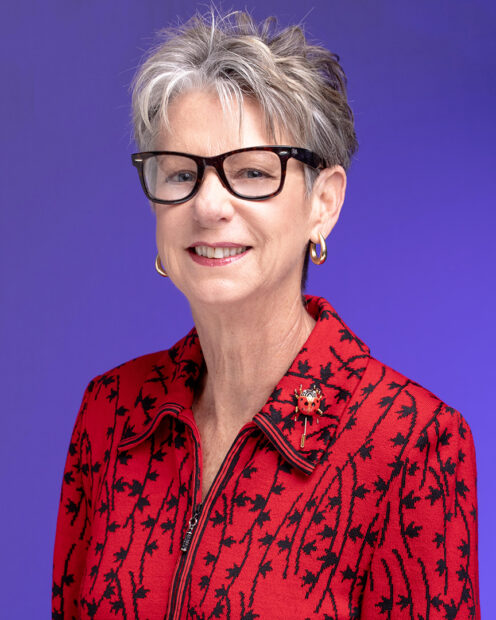 A headshot of Paulette Kluge. She wears a vibrant red collared shirt and is set against a deep purple background.