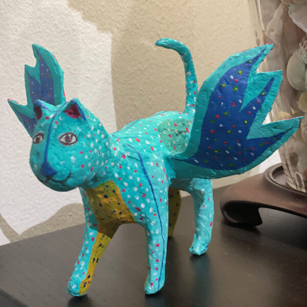 A photograph of a hand-crated alebrije. The small sculpture is of a cat-like creature with wings and is painted a bright shade of blue.