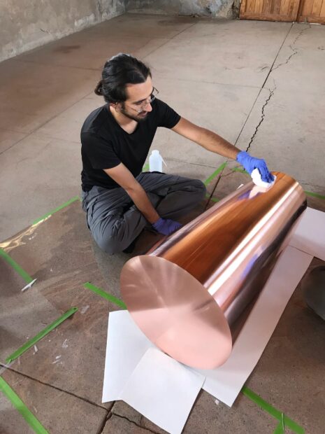 A photograph of an intern seated on the ground and cleaning a metal cylinder artwork.