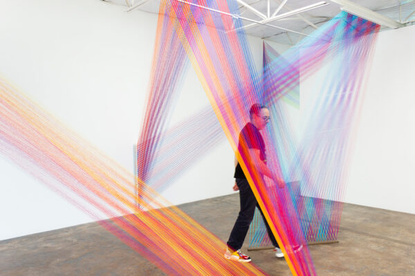 A photograph of artist Gabriel Dawe working on a colorful site-specific installation in a white walled gallery.