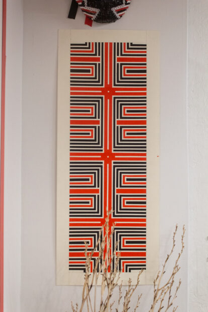 Red and black paint is applied in sharp right angles to a long rectangular piece of paper and hung on the wall.
