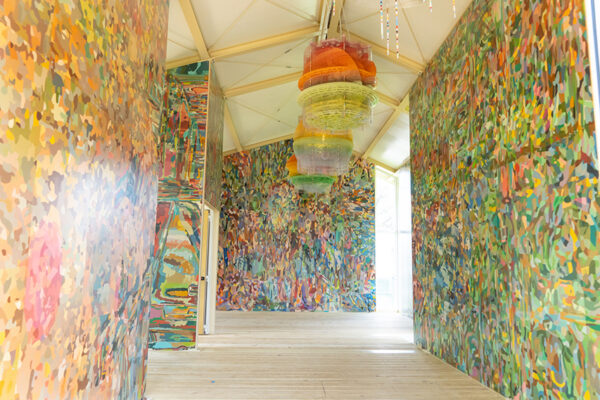 A photograph of the interior of a public art work by Jorge Pardo. The inside of this building is filled with colorful patterned paintings on the wall and has three vibrant and whimsical sculptures hanging from the ceiling.