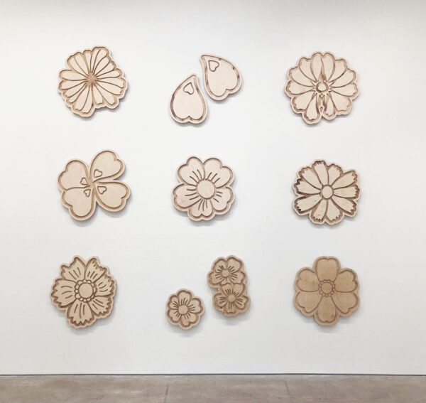A photograph of a grid of flowers carved from birch wood panels installed on a white gallery wall.