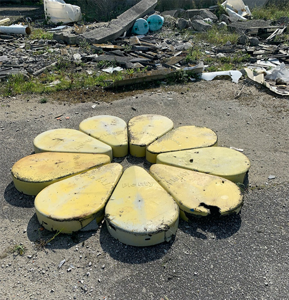 A photograph of a work by Sean Donovan. The image appears to be taken near a landfill and features nine yellow metal pieces arranged to look like flower petals.