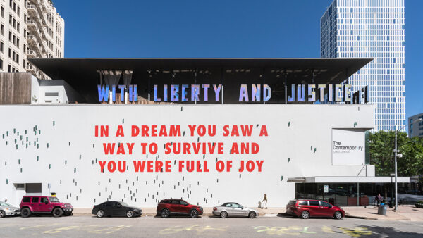 A photograph of a large-scale mural featuring all capitalized red letters on a white background. The text reads, "IN A DREAM YOU SAW A WAY TO SURVIVE AND YOU WERE FULL OF JOY."