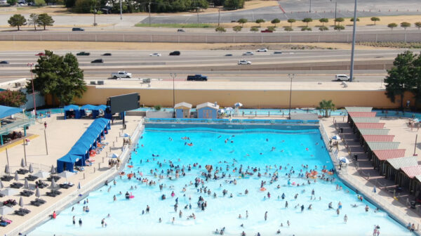 A still image from a video showing a large crowd of people in a wave pool that is set near a highway.