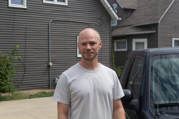William Anderson stands outside of his studio in a gray t-shirt. His head is shaved, and he is standing next to his black truck.