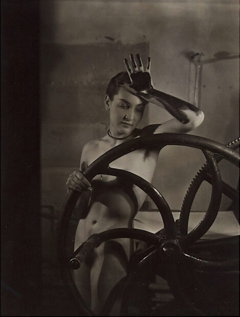 Nude female leaning against a press with her inked hand on her head