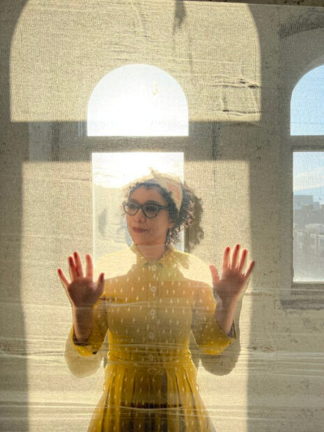 A photograph of artist Rehab El Sadek standing behind a translucent fabric with arched windows behind her.