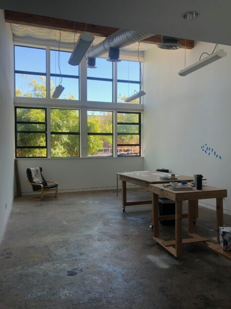 A white studio space with large glass windows and concrete floors is mostly empty except for a couple of work benches.